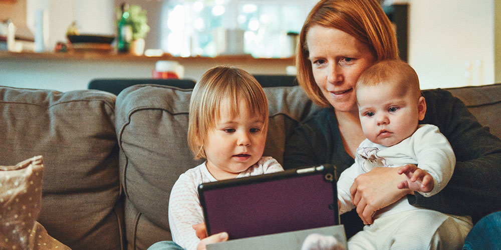 Woman holding a baby and toddler looking at a tablet sitting on the couch